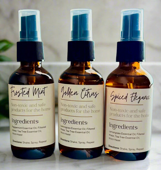 Three bottles of non-toxic bathroom spray with labels Frosted Mint, Golden Citrus, and Spiced Elegance, each with a spray nozzle and ingredient list on the label.